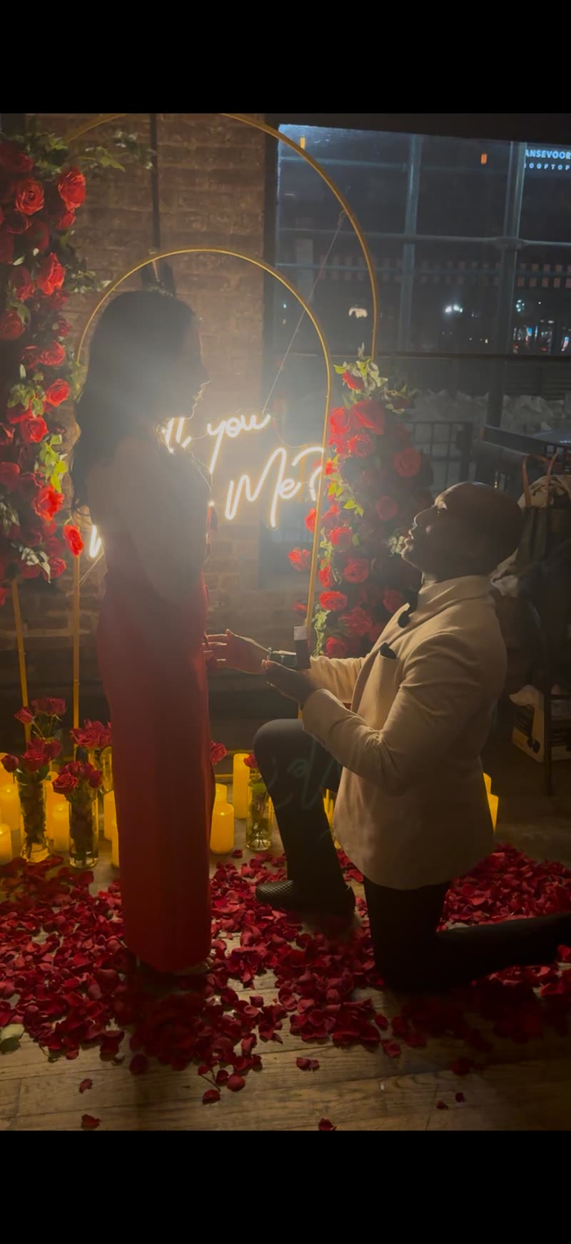 André on one knee proposing to Alexandra under a neon sign
